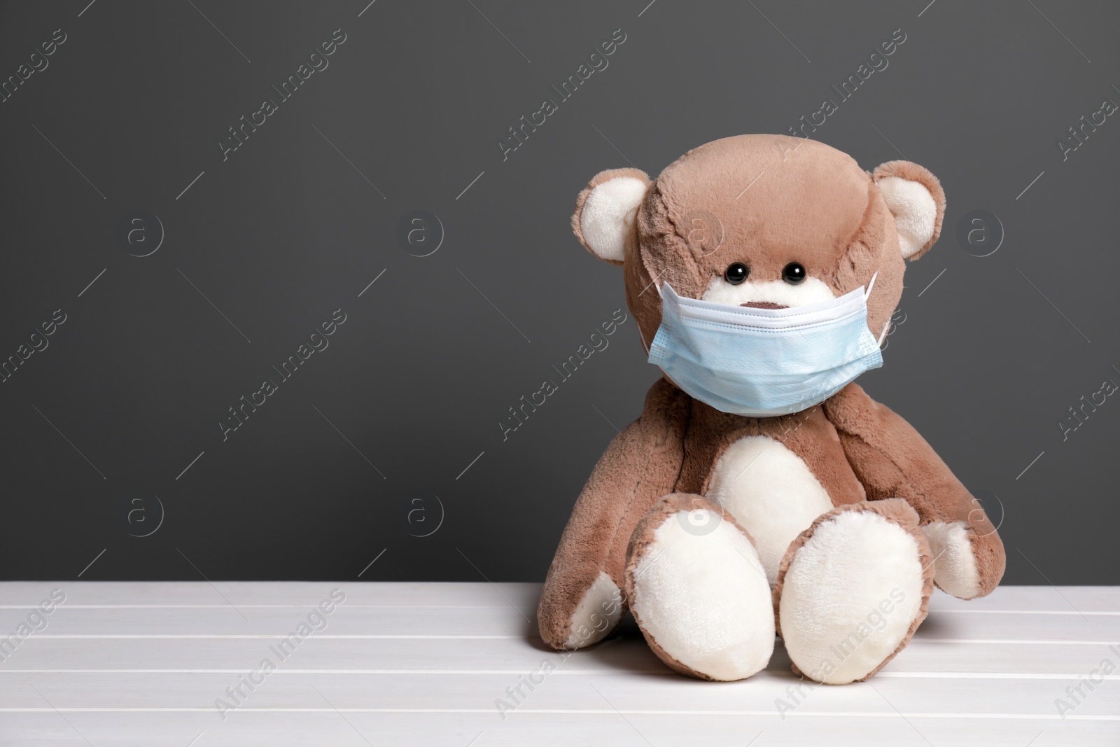 Photo of Cute teddy bear in medical mask on white wooden table near black wall, space for text