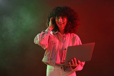 Photo of Beautiful young woman with laptop on color background in neon lights and smoke