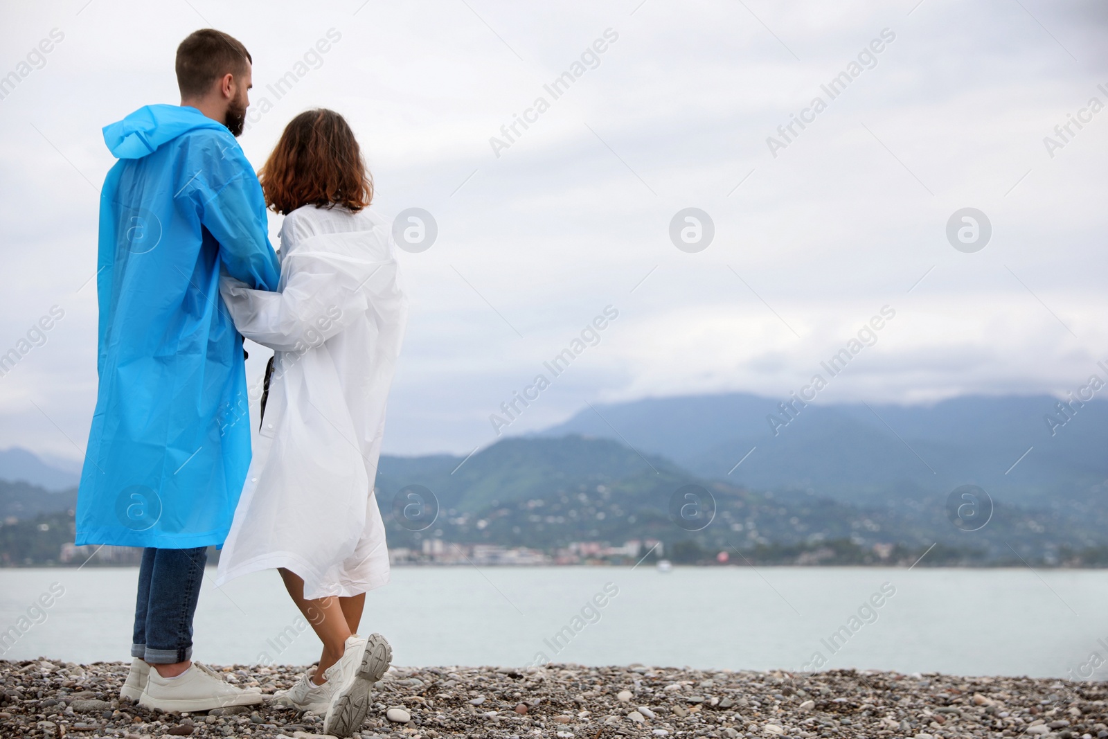 Photo of Young couple in raincoats enjoying time together under rain on beach, space for text