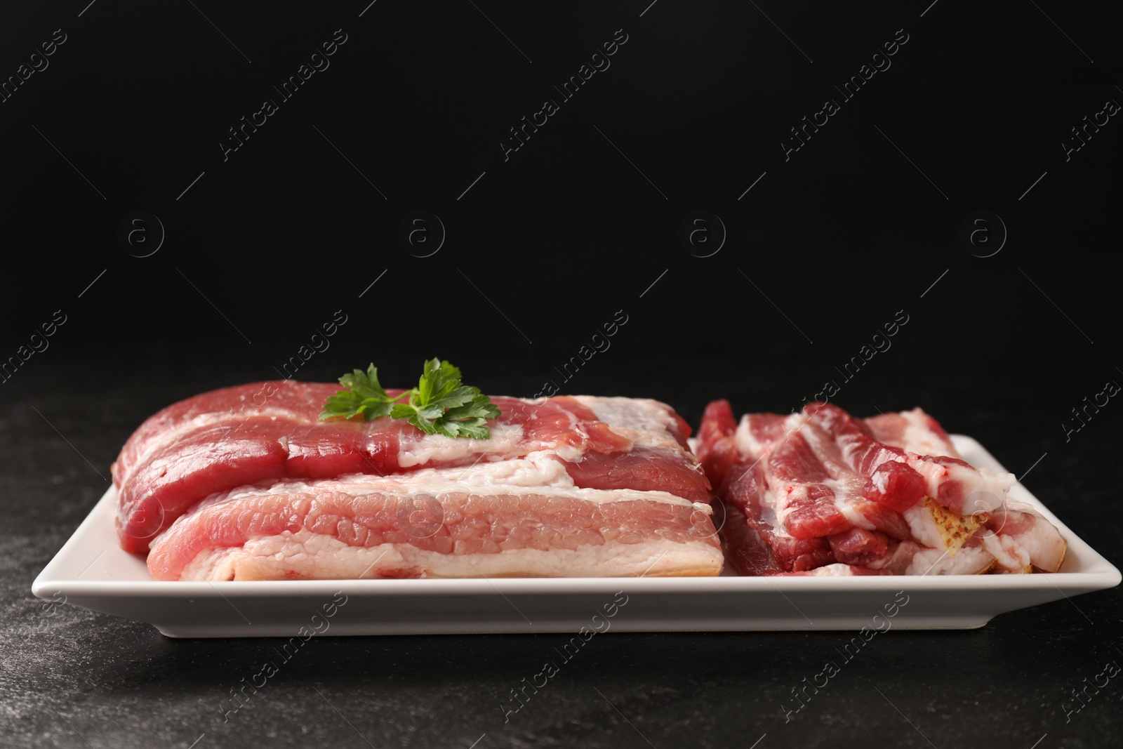 Photo of Pieces of raw pork belly and parsley on black textured table, closeup. Space for text