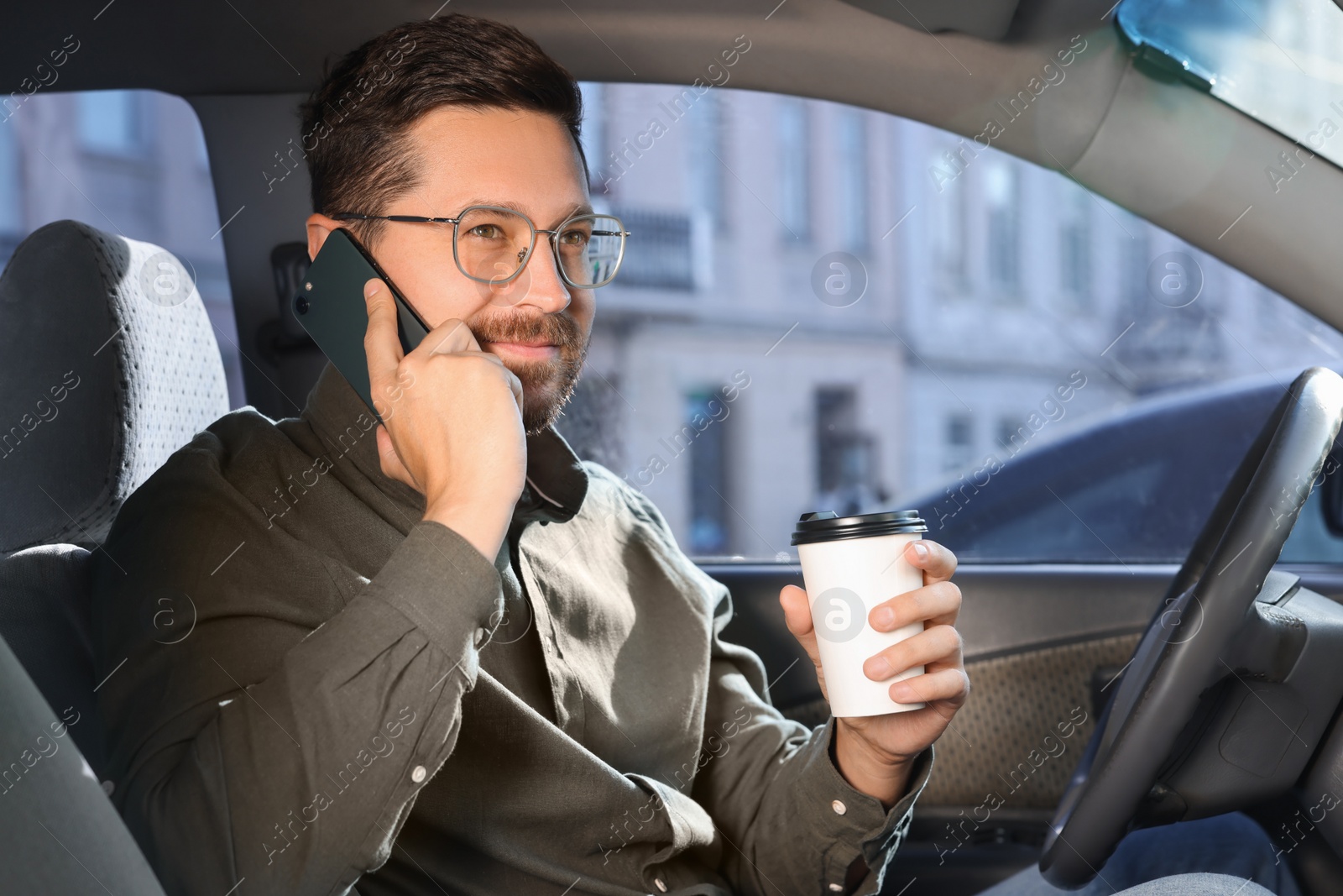 Photo of Coffee to go. Smiling man with paper cup of drink talking on smartphone in car