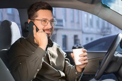 Photo of Coffee to go. Smiling man with paper cup of drink talking on smartphone in car