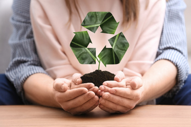 Image of Couple holding fertile soil at wooden table and recycling symbol, closeup