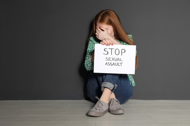 Photo of Young woman holding card with words STOP SEXUAL ASSAULT while sitting near grey wall. Space for text