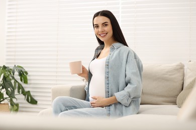 Happy pregnant woman with cup of drink on sofa at home