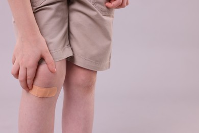 Little boy putting sticking plaster onto knee against light grey background, closeup. Space for text