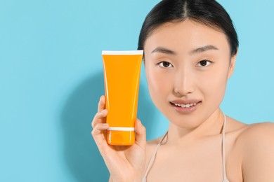 Beautiful young woman holding sun protection cream on light blue background, space for text