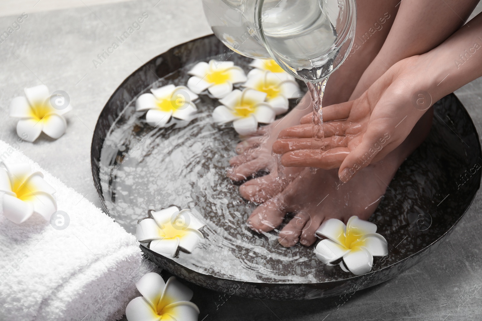 Photo of Woman pouring water onto hand while soaking her feet in bowl on light grey floor, closeup. Spa treatment