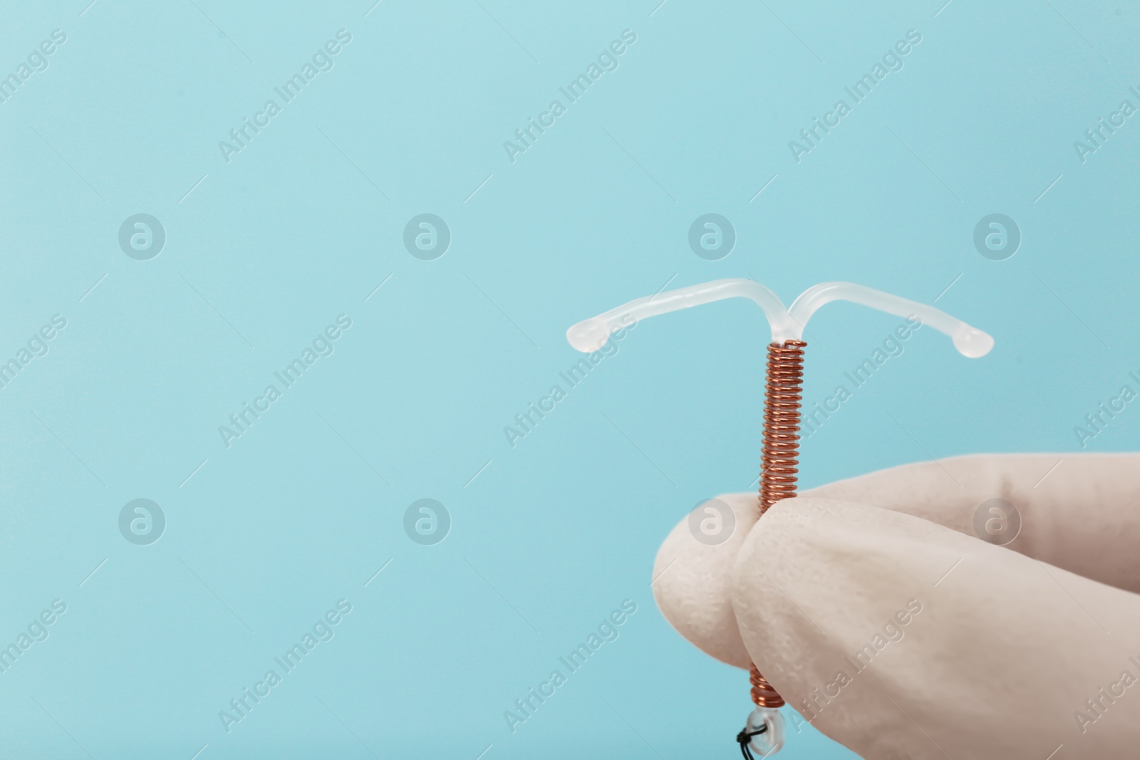 Photo of Doctor holding T-shaped intrauterine birth control device on light blue background, closeup. Space for text