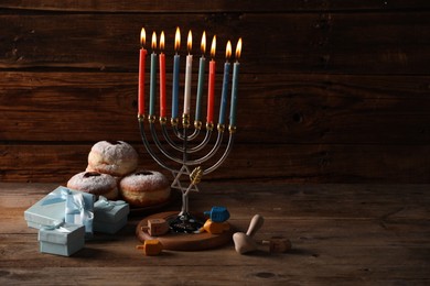 Photo of Hanukkah celebration. Menorah with burning candles, dreidels, donuts and gift boxes on wooden table, space for text