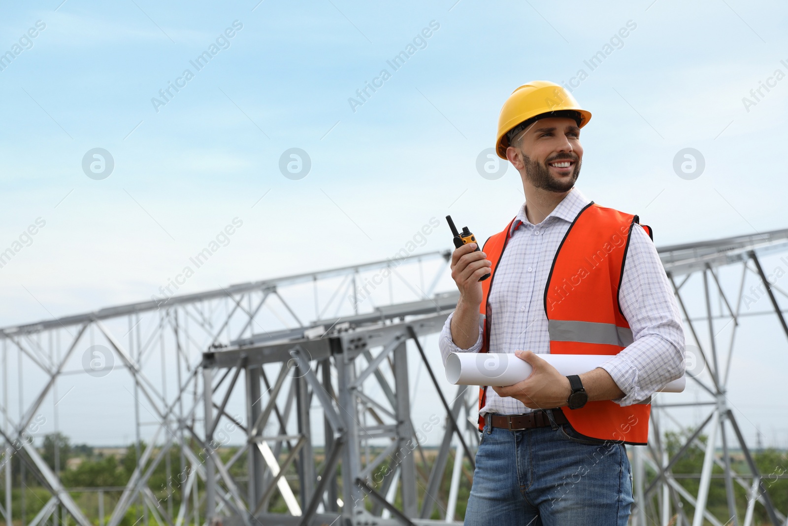 Photo of Professional engineer with plan and walkie talkie near high voltage tower construction outdoors. Installation of electrical substation