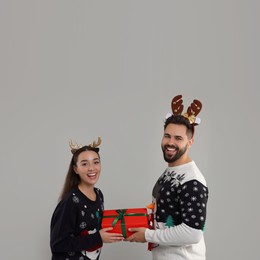 Happy young couple in Christmas sweaters and reindeer headbands holding gift on grey background