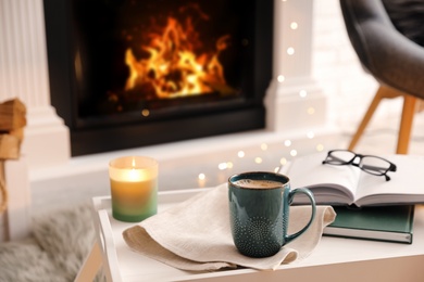 Photo of Cup of coffee, burning candle and books on tray near fireplace indoors. Cozy atmosphere