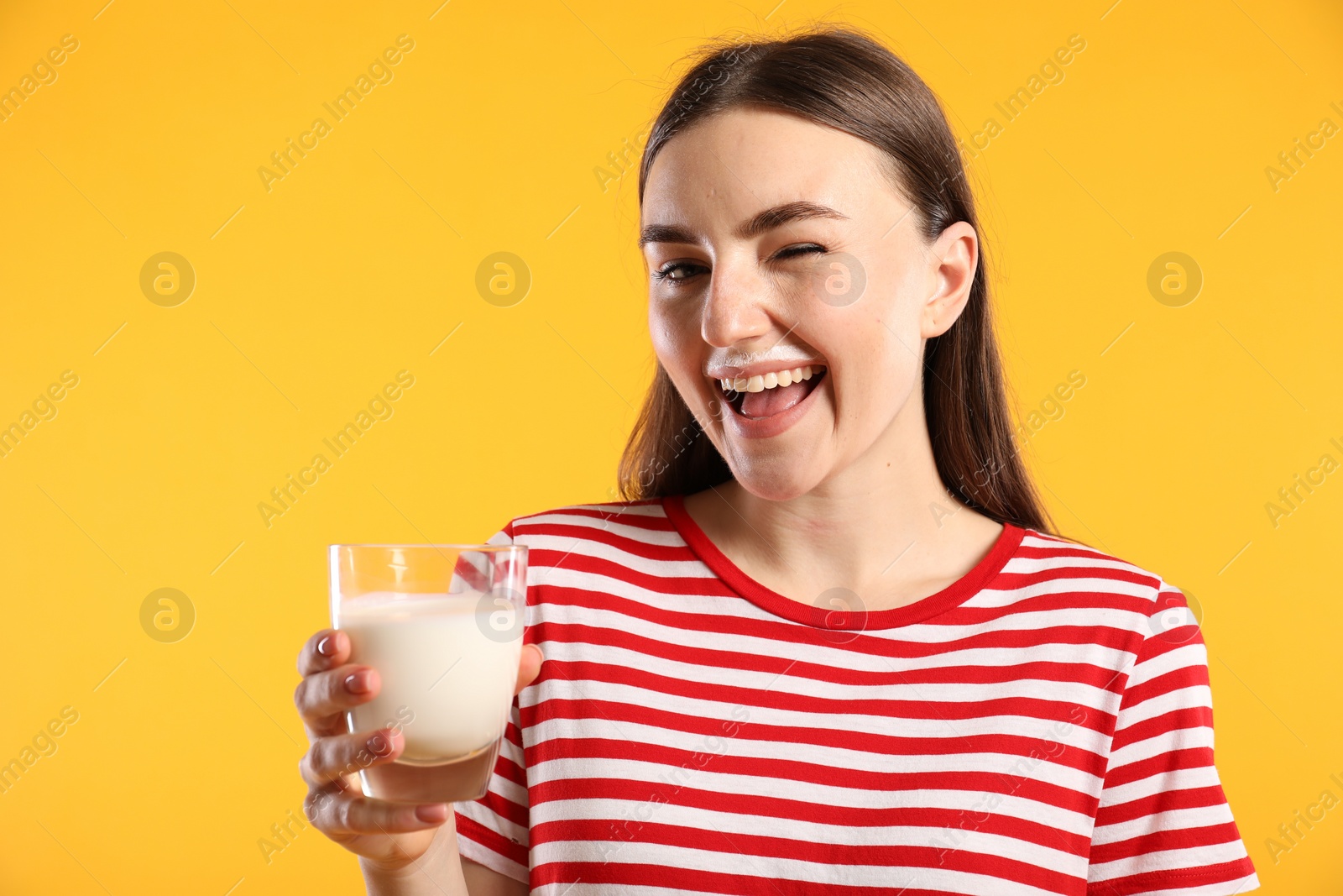 Photo of Happy woman with milk mustache holding glass of tasty dairy drink and winking on orange background