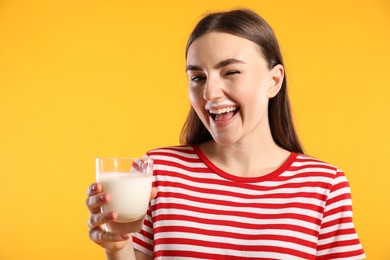 Photo of Happy woman with milk mustache holding glass of tasty dairy drink and winking on orange background