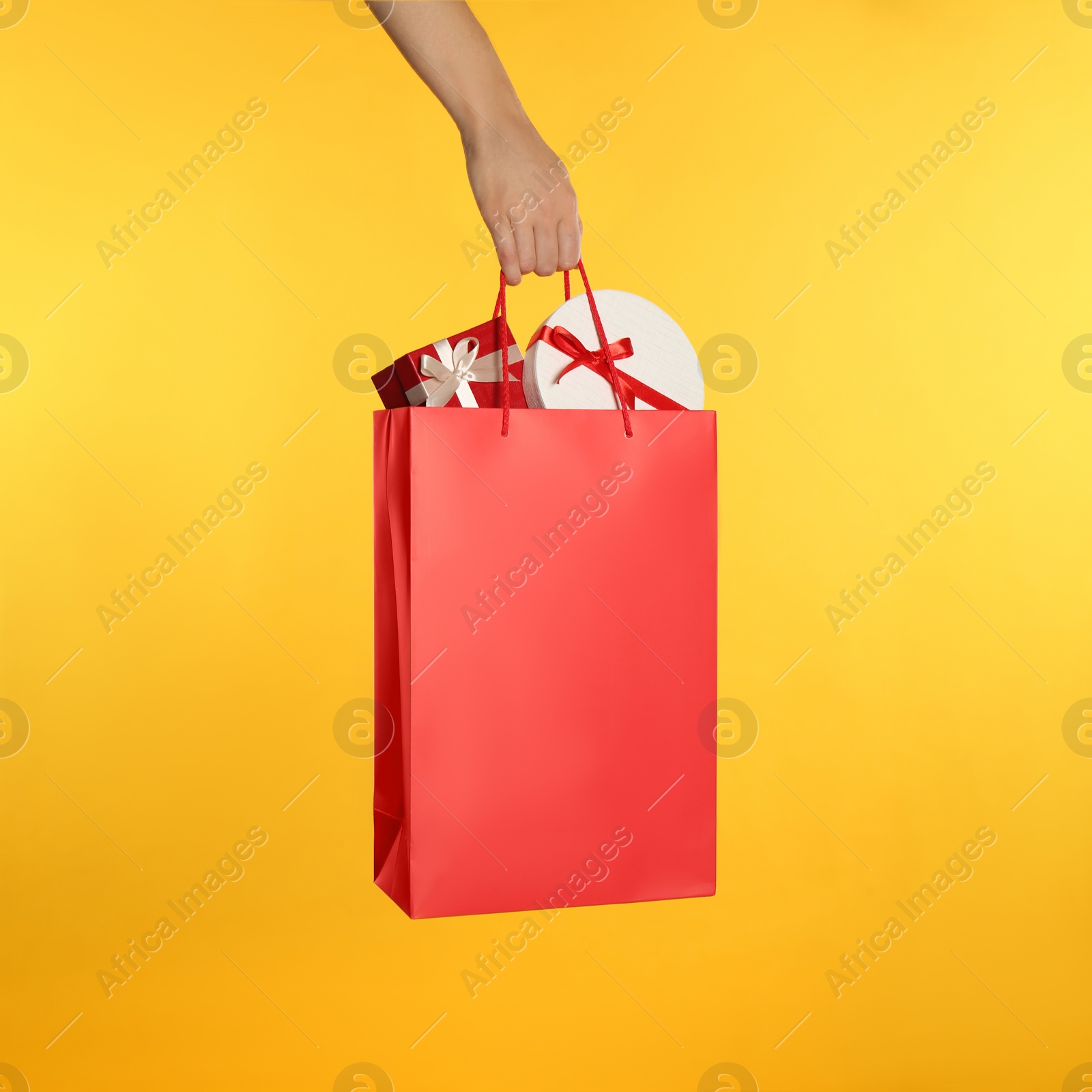 Photo of Woman holding paper shopping bag full of gift boxes on yellow background, closeup