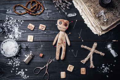 Voodoo doll with pins surrounded by ceremonial items on black wooden table, flat lay