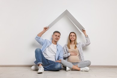 Young family housing concept. Pregnant woman with her husband sitting under cardboard roof on floor indoors