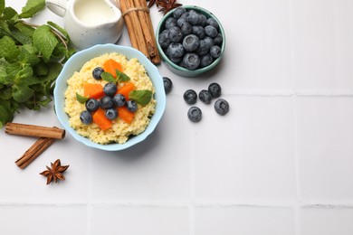 Photo of Tasty millet porridge with blueberries, pumpkin and mint in bowl on white tiled table, flat lay. Space for text