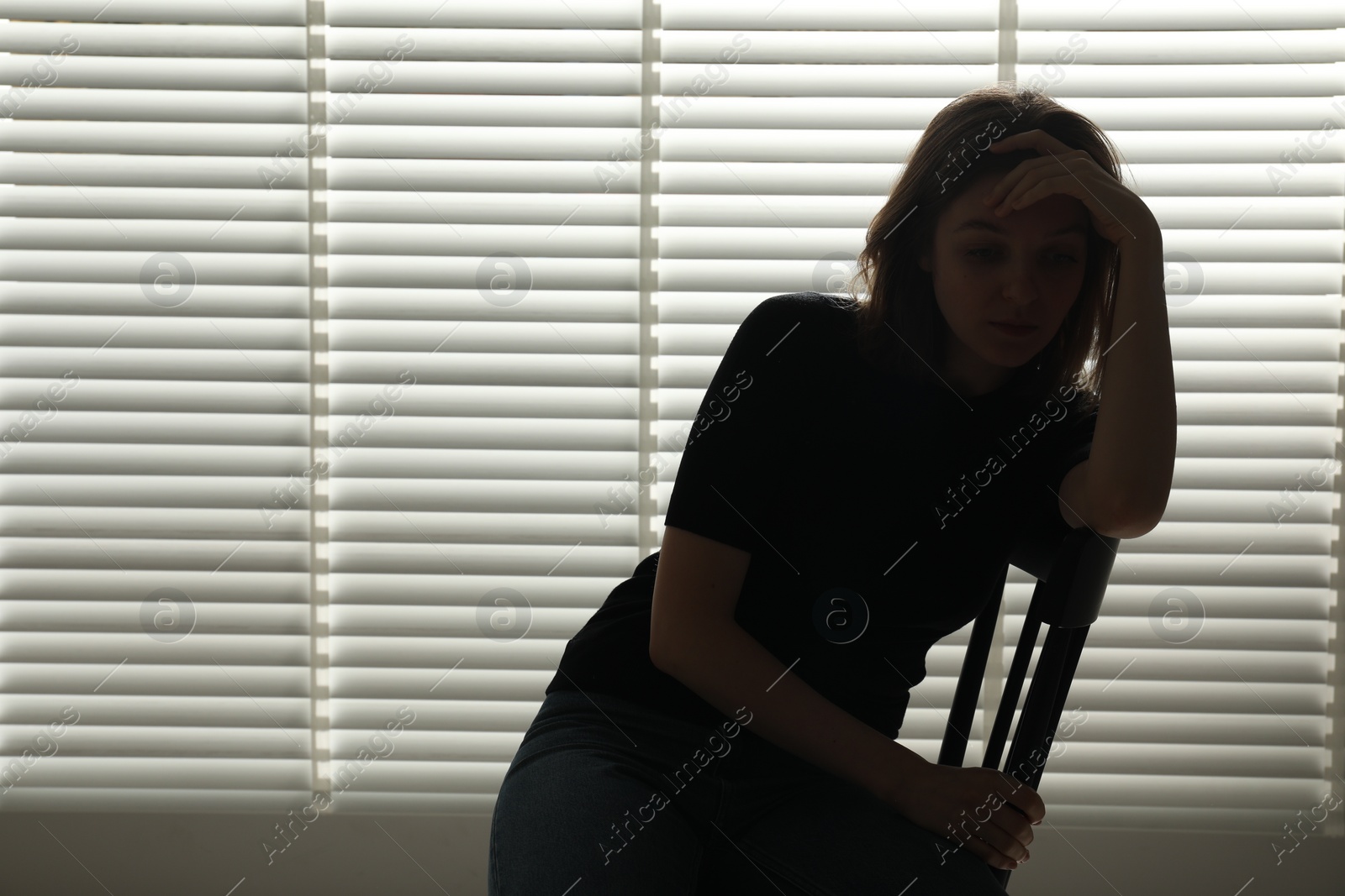 Photo of Silhouette of sad young woman near closed blinds indoors, space for text