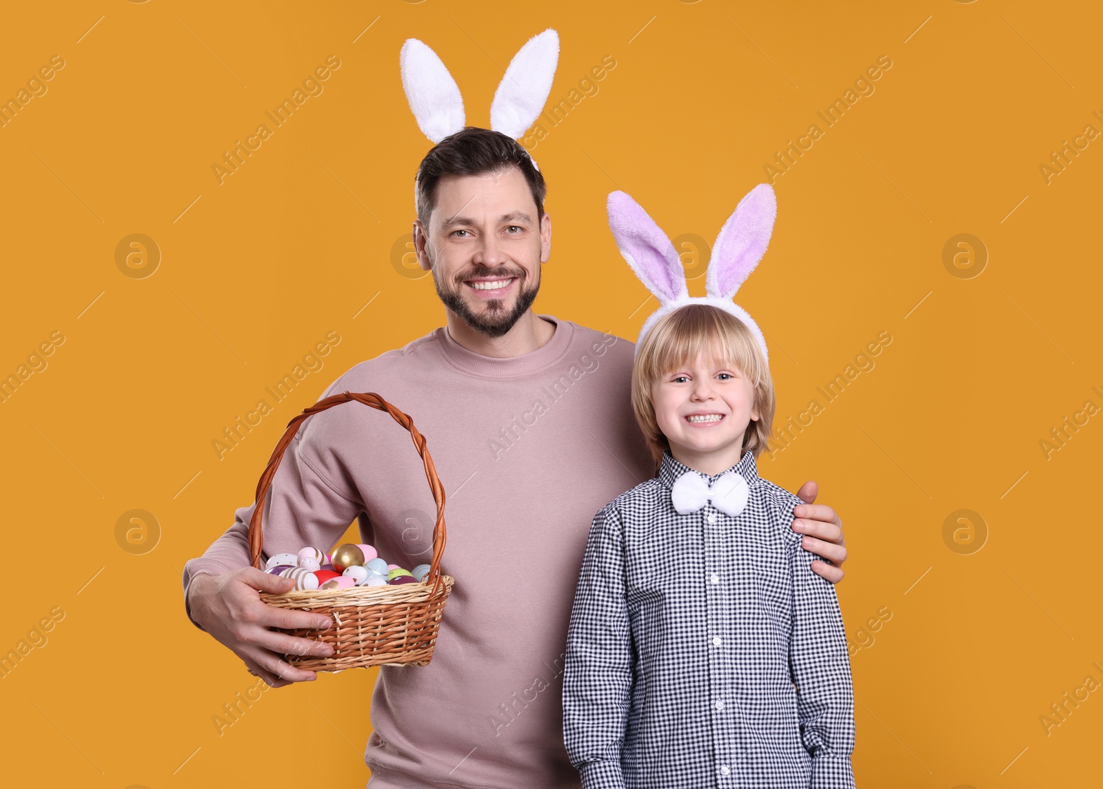 Photo of Father and son in bunny ears headbands with wicker basket of painted Easter eggs on orange background