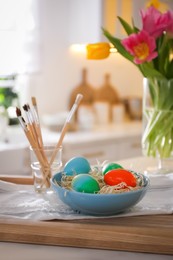 Beautifully painted Easter eggs in bowl and brushes on table indoors