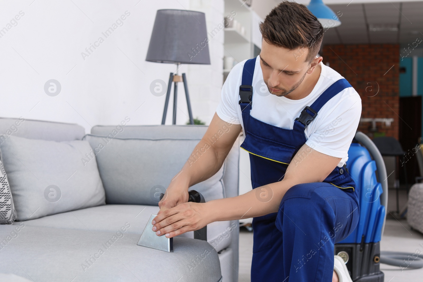 Photo of Male worker removing dirt from sofa with professional vacuum cleaner indoors