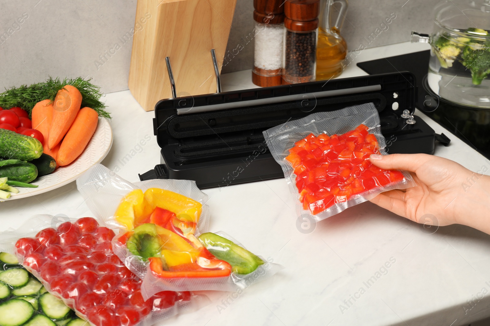 Photo of Woman using sealer for vacuum packing with plastic bag of red pepper at white table, closeup