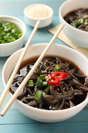 Photo of Tasty soup with buckwheat noodles (soba), chili pepper, green onion in bowl and chopsticks on light blue wooden table, closeup