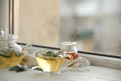 Cup of sage tea and green leaves on white wooden windowsill. Space for text