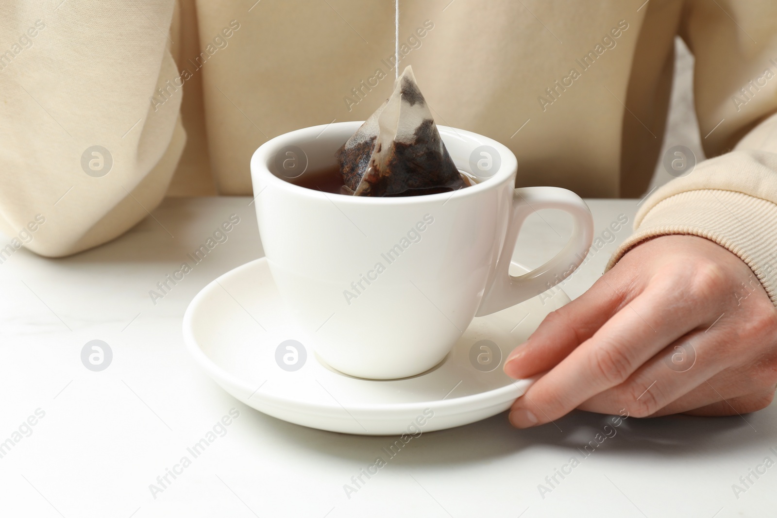 Photo of Woman putting tea bag in cup with hot water at white table, closeup