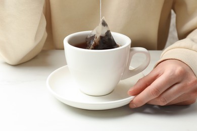 Woman putting tea bag in cup with hot water at white table, closeup