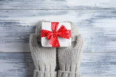 Photo of Woman wearing warm mittens holding Christmas gift on wooden background, top view