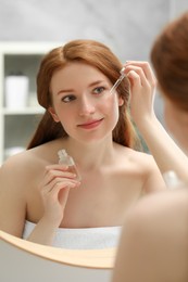 Beautiful woman with freckles applying cosmetic serum onto her face near mirror in bathroom