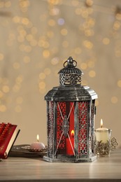 Arabic lantern, Quran, misbaha and candles on wooden table against blurred lights