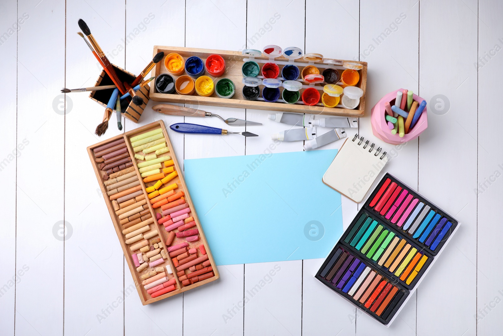 Photo of Blank sheet of paper, colorful chalk pastels and other drawing tools on white wooden table, flat lay. Modern artist's workplace