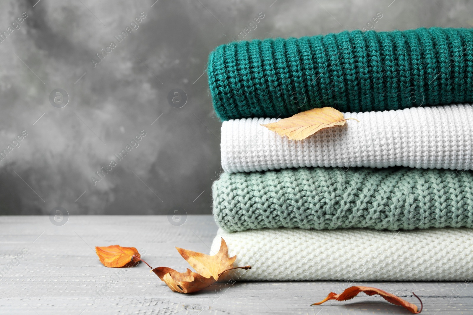 Photo of Stack of warm clothes and autumn leaves on wooden table against grey background. Space for text