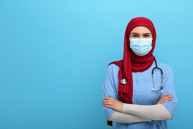 Photo of Muslim woman wearing hijab, medical uniform and protective mask on light blue background, space for text