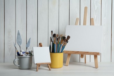 Photo of Easels with blank canvases, paints and brushes on white wooden table
