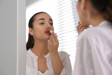 Photo of Young woman with herpes applying lip balm in front of mirror at home