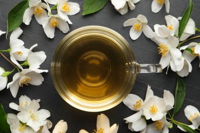 Photo of Glass cup of jasmine tea and fresh flowers on black table, flat lay