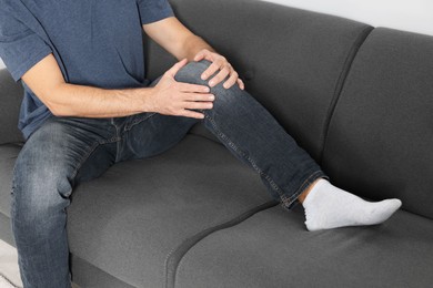 Photo of Man suffering from leg pain and touching knee on sofa, closeup