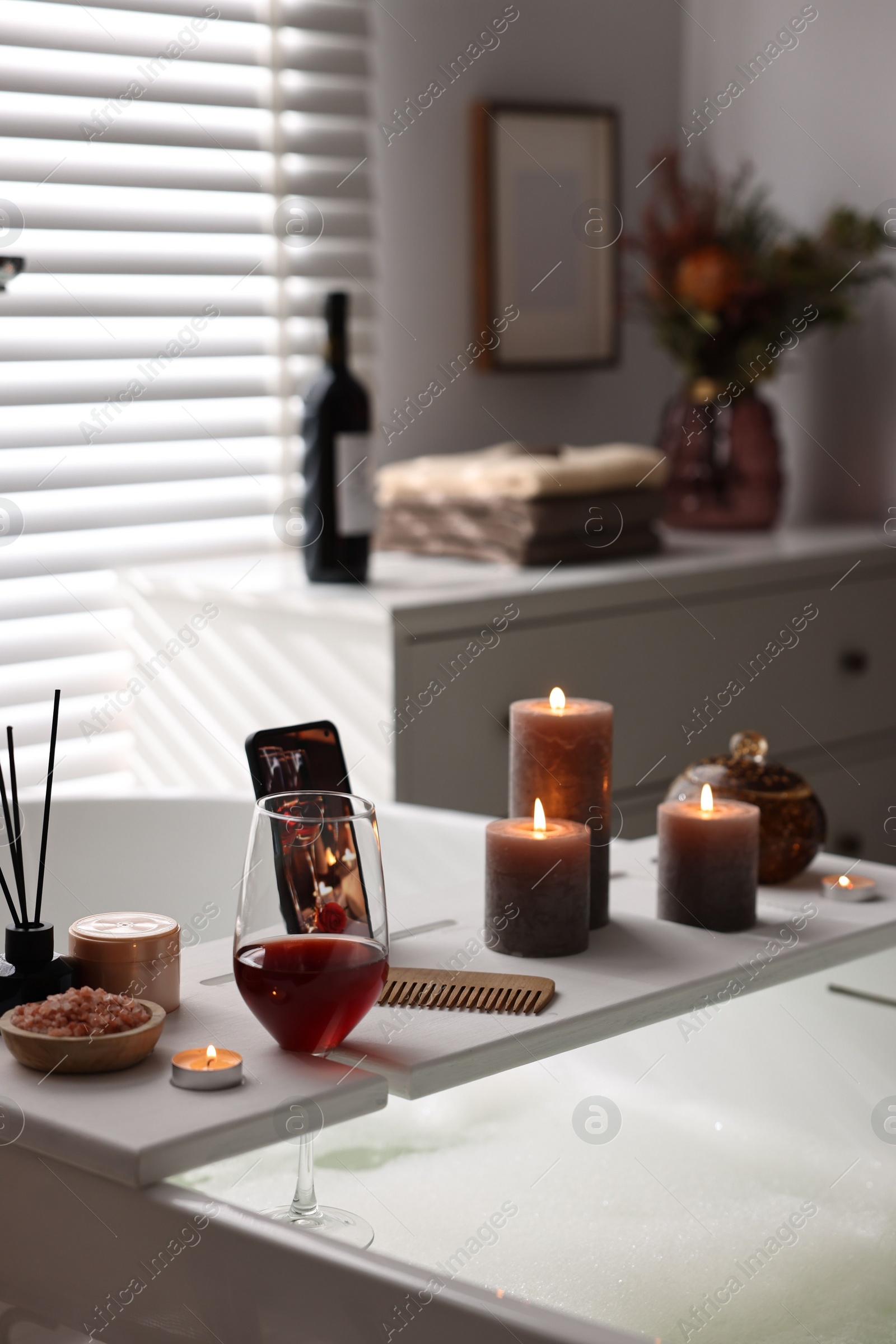 Photo of White wooden tray with glass of wine, beauty products and burning candles on bathtub in bathroom