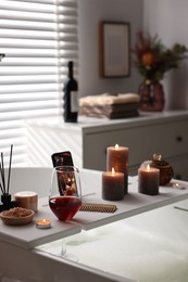 Photo of White wooden tray with glass of wine, beauty products and burning candles on bathtub in bathroom