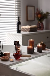 White wooden tray with glass of wine, beauty products and burning candles on bathtub in bathroom