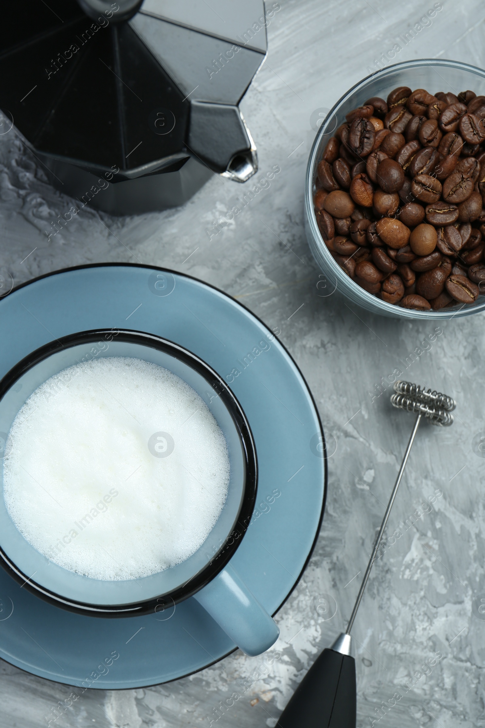 Photo of Mini mixer (milk frother), whipped milk in cup, coffee beans and moka pot on grey textured table, flat lay