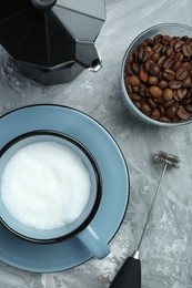 Photo of Mini mixer (milk frother), whipped milk in cup, coffee beans and moka pot on grey textured table, flat lay