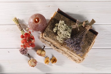 Different dry herbs, flowers, old book and burning candle on white wooden table, flat lay