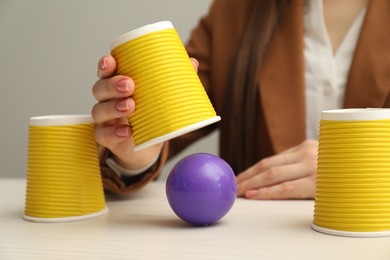 Shell game. Woman showing ball under cup at white wooden table, closeup
