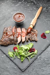 Photo of Pieces of delicious roasted beef meat with sauce and greens on black table